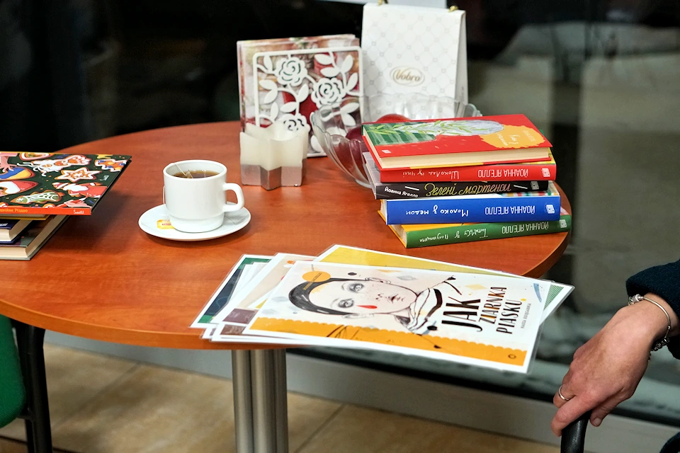 Na okrągłym stoliku leży kilka książek, filiżanka kawy, serwetki, świeczka. Leżą też zafoliowane plakaty formatu A3. Ten na wierzchu prezentuje rysunek portretowy dziewczynki i tytuł: Jak ziarnka piasku.