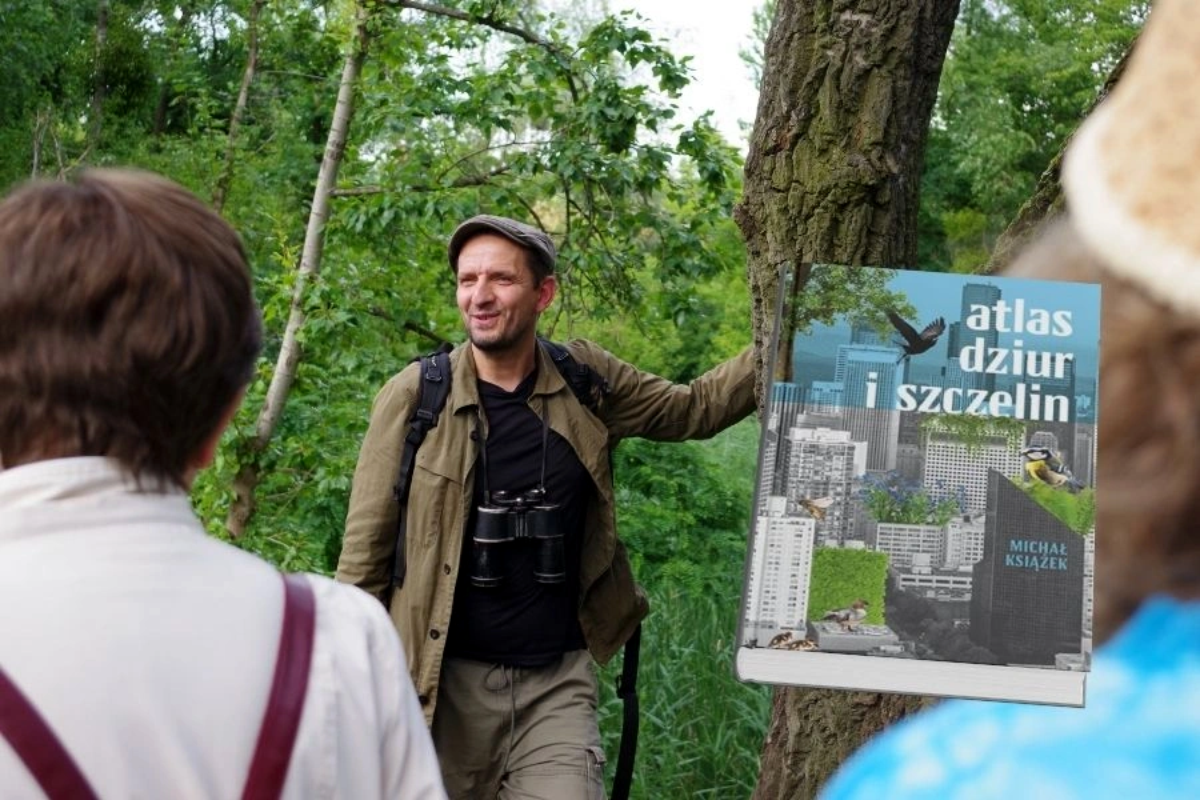 Osoba w zielonej kurtce z lornetką wskazująca na książkę zatytułowaną „Atlas dzikiej i szczęśliwej” wyeksponowaną na tle drzewa w bujnym lesie.