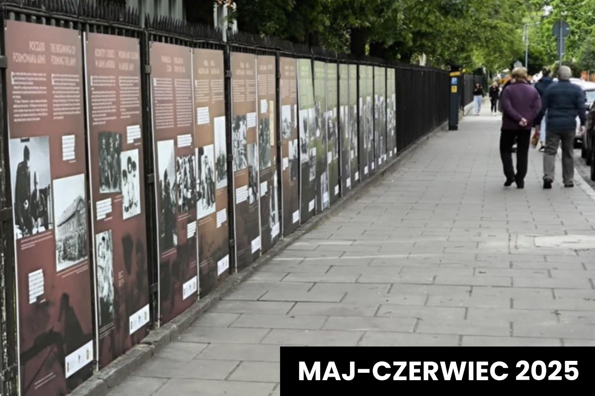 Plansze wystawowe wiszą na płocie, dwóch mężczyzn idzie chodnikiem