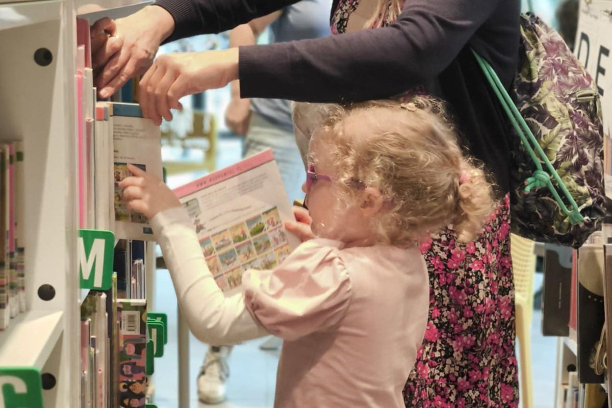 Dziewczynka z jasnymi włosami wkłada książkę na półkę w bibliotece, w tle jej mama