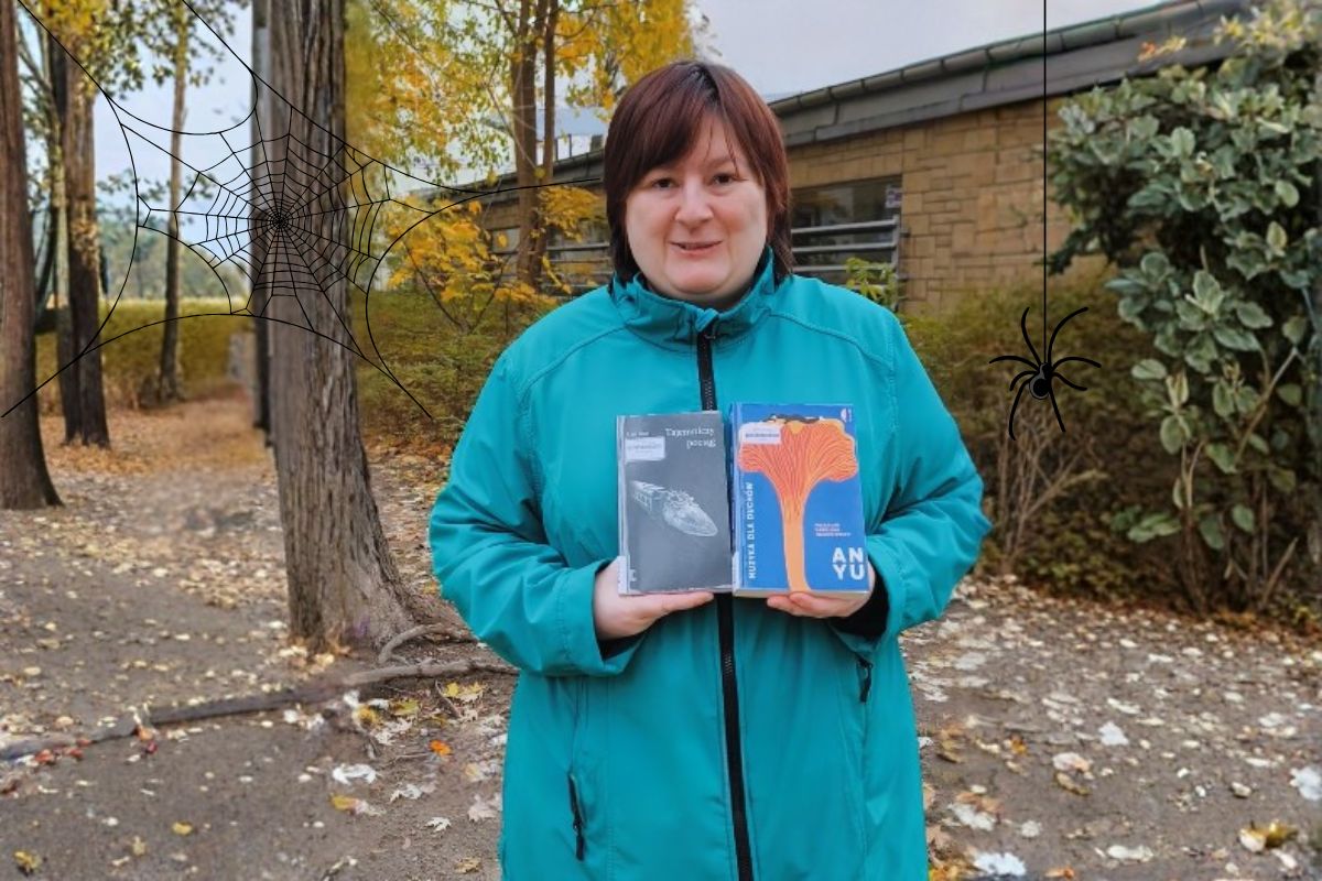 Kobieta w turkusowej kurtce trzyma w ręku książki, w tle budynek i jesienne drzewa