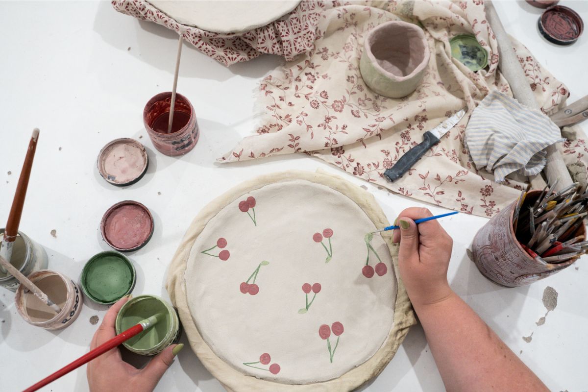 Ręce kobiety malujące gliniany talerz w wisienki, słoiczki z farbami, gliniane naczynia, serweta w kwiaty, kubek z pędzlami