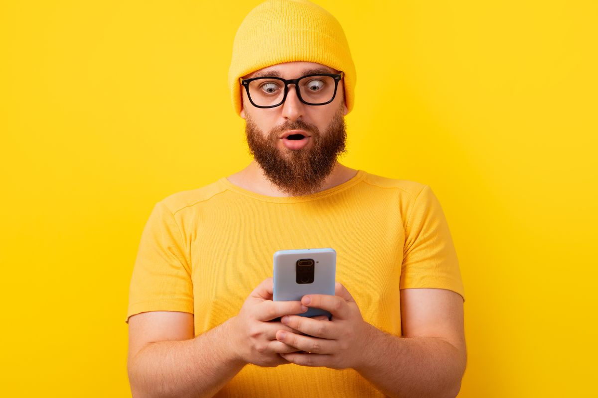 Na żótym tle brodaty mężczyzna w żółtej czapce i tiszercie ze zdziwieniem patrzy w telefon