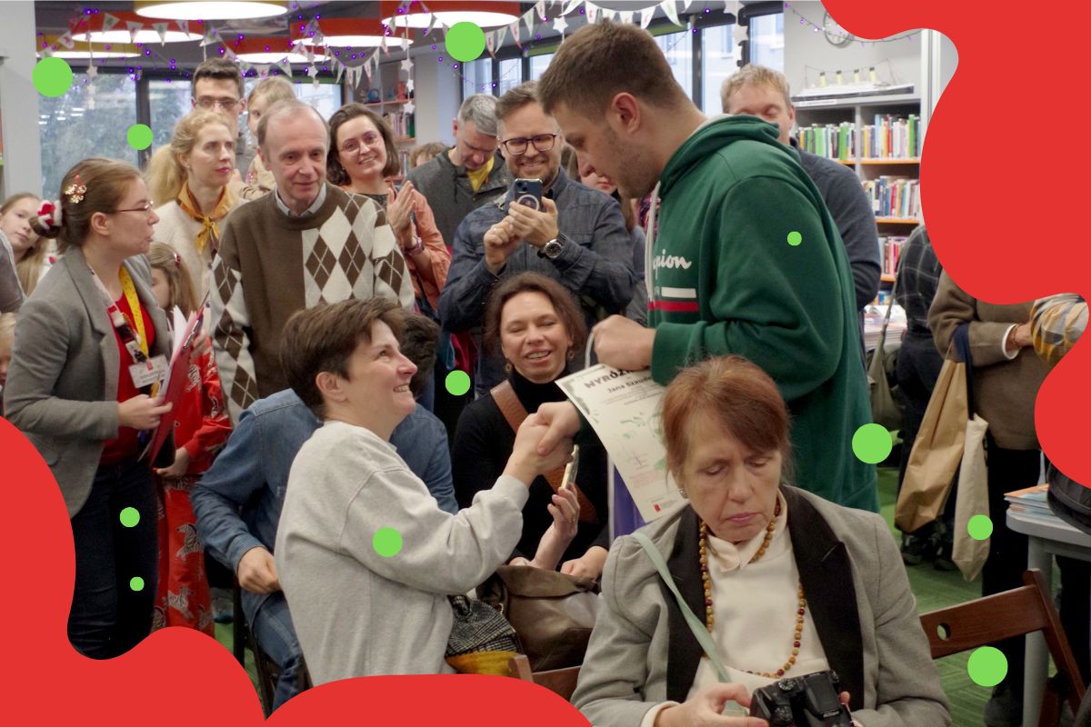 Grupa uśmiechnietych ludzi, mężczyzna trzyma dyplom i gratuluje kobiecie, mężczyzna robi zdjęcie smartfonem