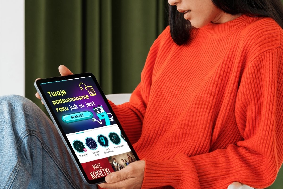 Kobieta w pomarańczowym swetrze trzyma w ręku tablet i patrzy na niego