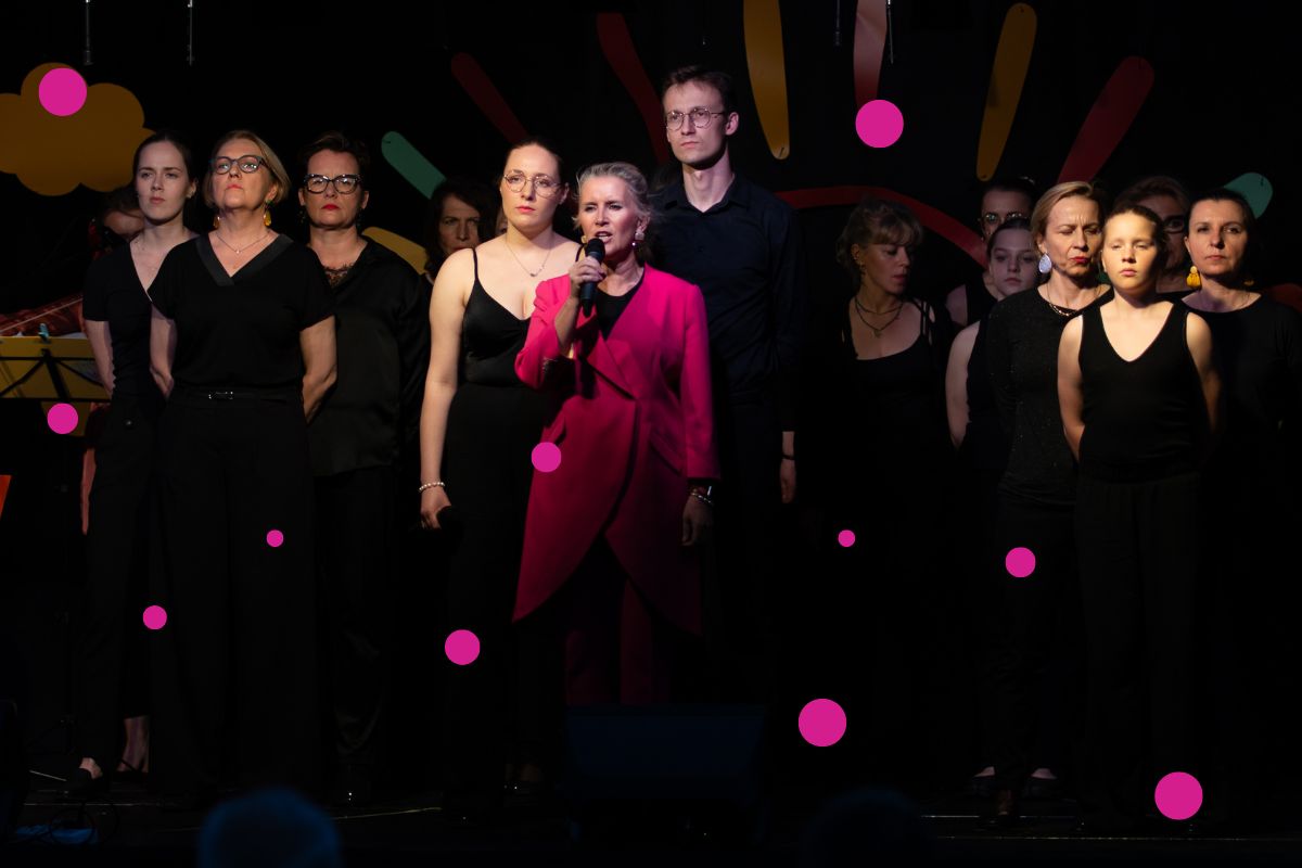Grupa kobiet i mężczyzna w czarnych ubraniach, kobieta w różowej sukni śpiewa, trzymając mikrofon w ręku