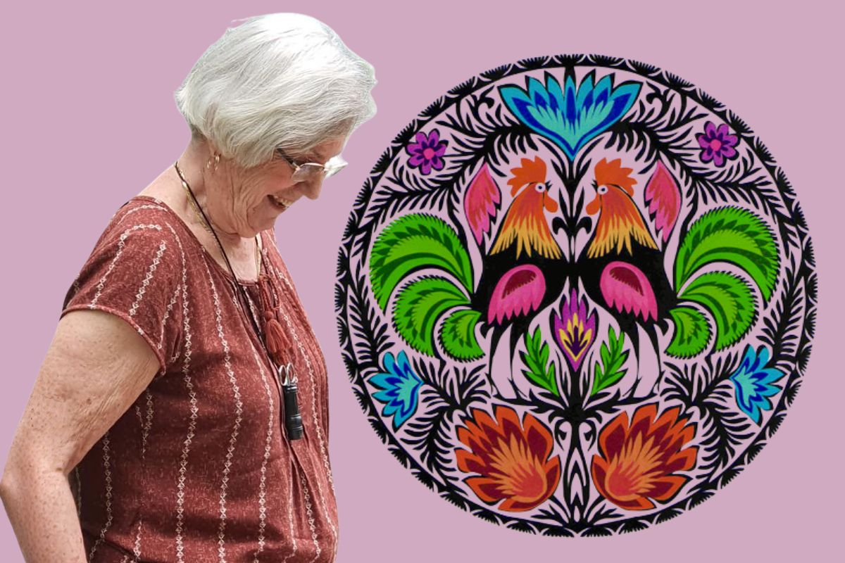 Na fioletowym tle kobieta z siwymi włosami i w okularach, obok wycinanka łowicka z kogutami