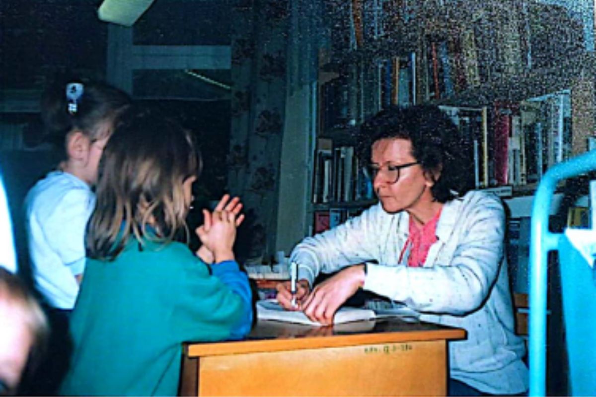 Przy bibliotecznej ladzie siedzi kobieta o ciemnych włosach, po drugiej stronie stoją dwie dziewczynki