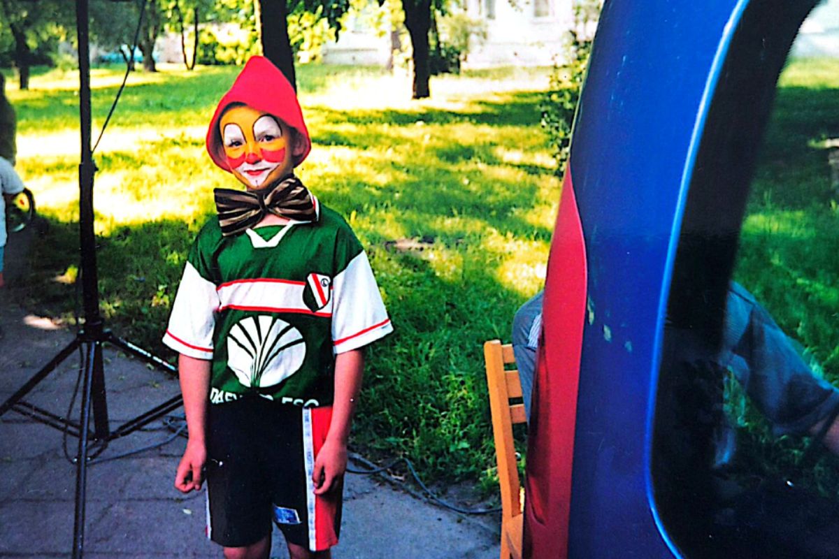 Chłopiec w czerwonej czapce z pomalowaną buzią jak klaun, w tle trawa i drzewa