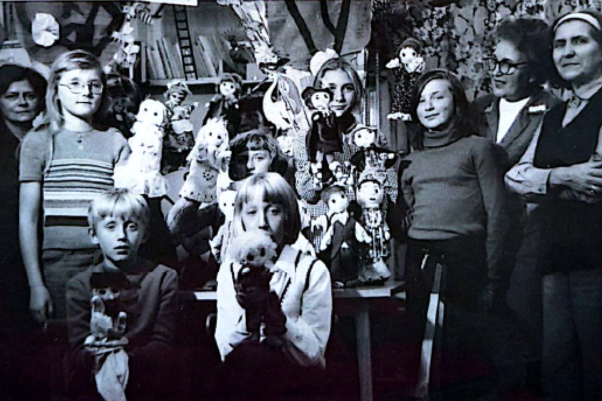 N czarno-białym zdjęciu grupa dorosłych i dzieci z kukiełkami teatralnymi