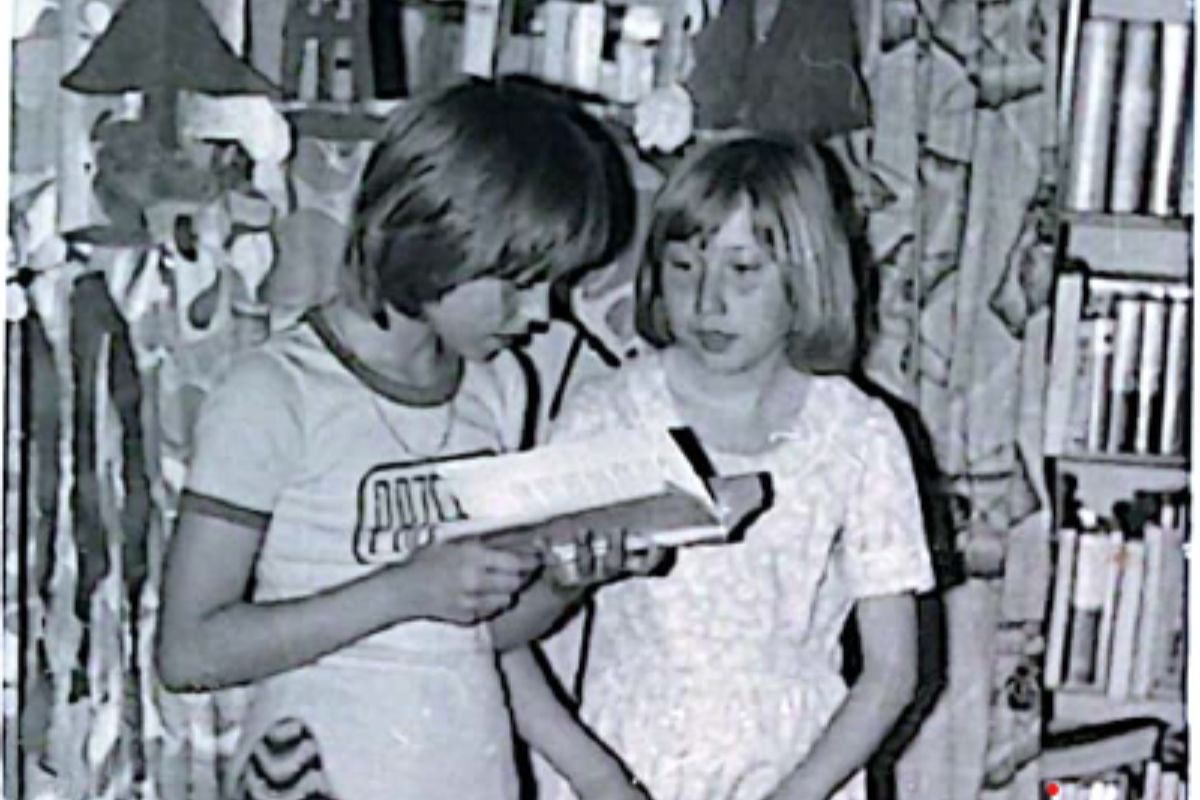 Dwie dziwczynki recytują wiersz, jedna czyta książkę, w tle zasłony i regały biblioteczne, zdjęcie czarno-białe
