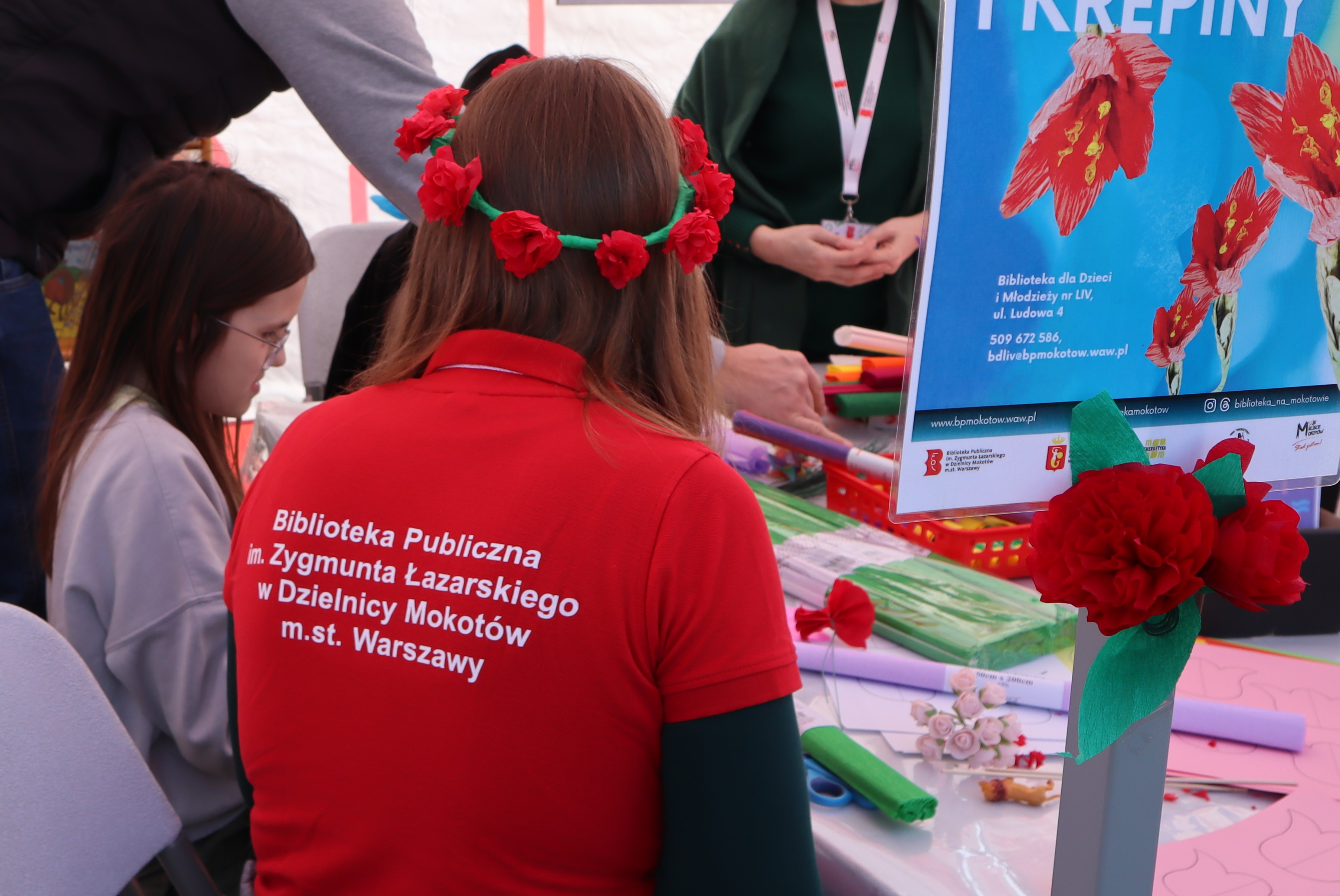 Kobieta w wianku z papieru i czerwonej koszulce bibliotecznej siedzi tyłem do obiektywu, z prawej papierowy kwiat i plakat, z lewej dziewczynka