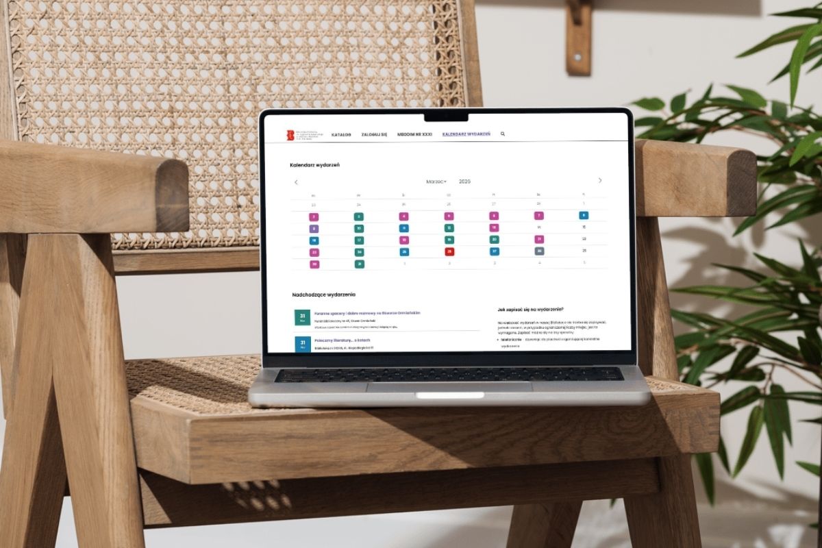 Na drewnianym krześle laptop z otwartą stroną Biblioteki Mokotów, z boku widoczne liście rośliny domowej