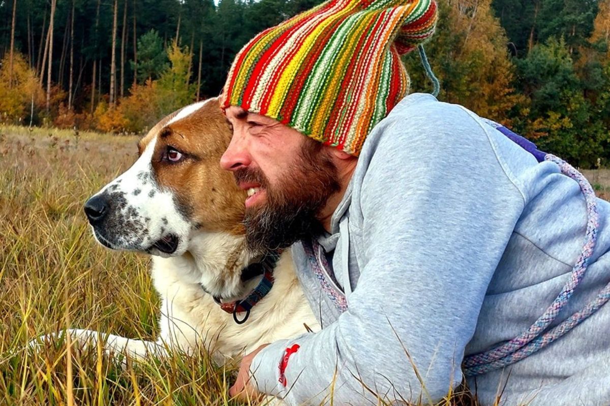 Mężczyzna z brodą i w kolorowej czapce leży razem ze swoim psem na łące