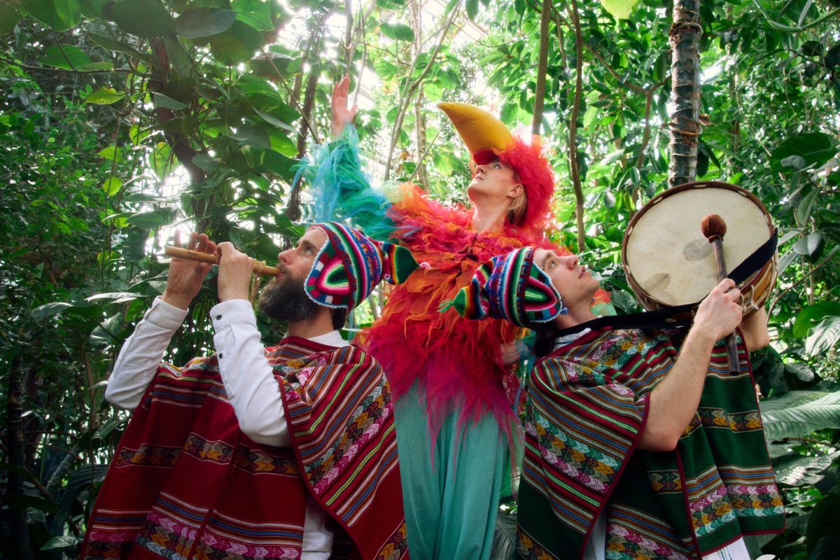 Na tle lasu troje ludzi: mężczyzna gra na fujarce, kobieta przebrana za papugę, mężczyzna gra na bębnie