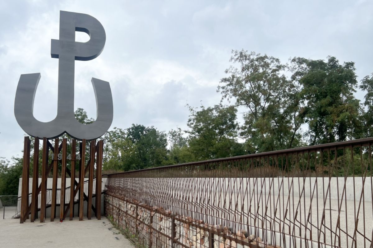 Na tle zachmurzonego nieba kotwica, znak Powstania Warszawskiego, metalowe ogrodzenie
