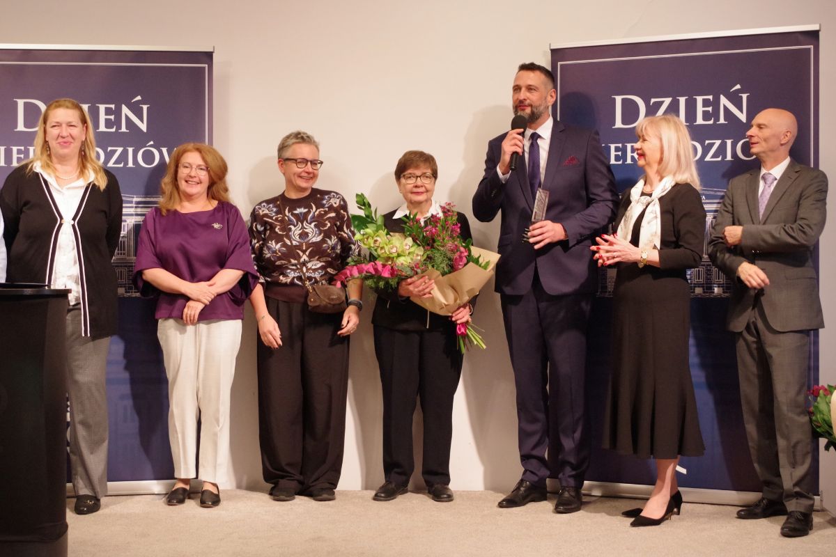 Grupa ludzi na tle standów z napisem: Dzień Kierbedziów, mężczyzna w garniturze mów do mikrofonu, kobieta trzyma kwiaty
