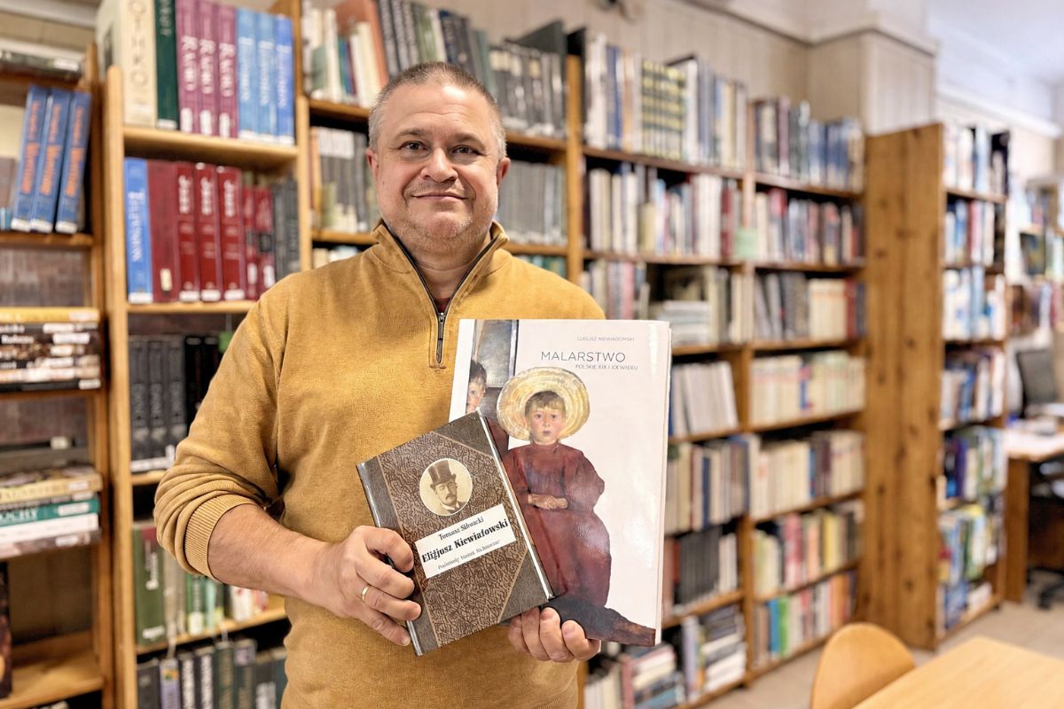 Mężczyzna w żółtym swetrze pokazuje dwie książki, w tle regały biblioteczne