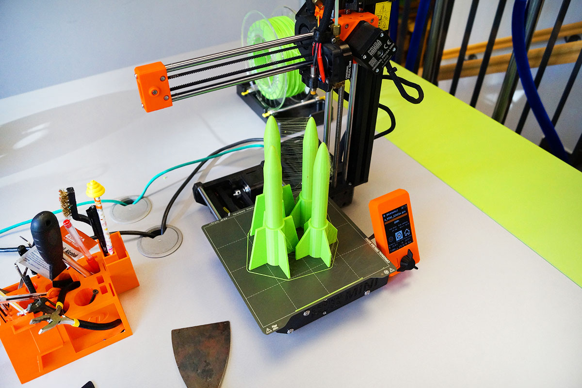 Drukarka 3D z czterema pionowo ustawionymi modelami rakiet kosmicznych na jej blacie.