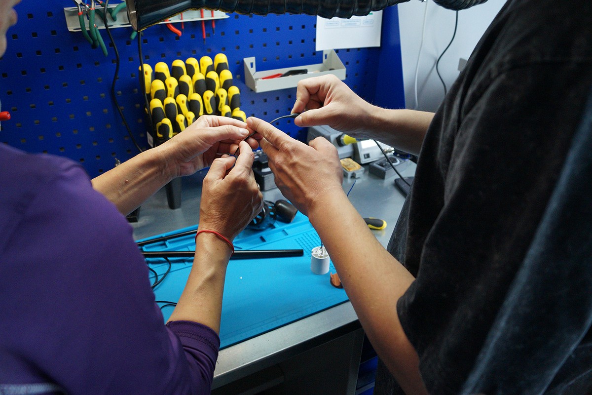 Dwie pary rąk trzymają kabel, a w tle widać warsztat z ułożonymi śrubokrętami