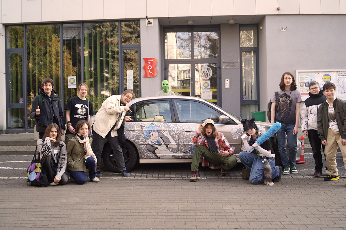 Grupa młodych osób pozuje przed samochodem, którego karoseria ozdobiona jest malunkiem w stylu mangowym