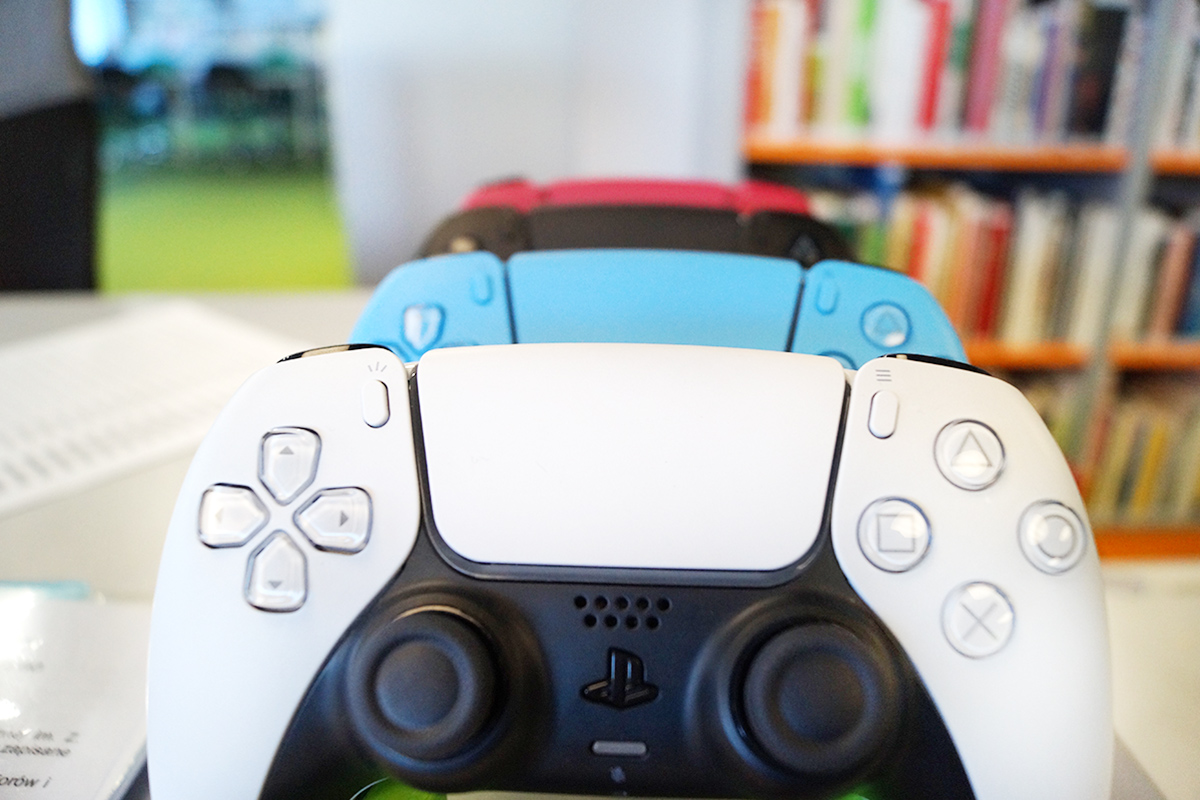 Ułożone w pionie, jeden za drugim kontrolery do Playstation, w tle regał z książkami