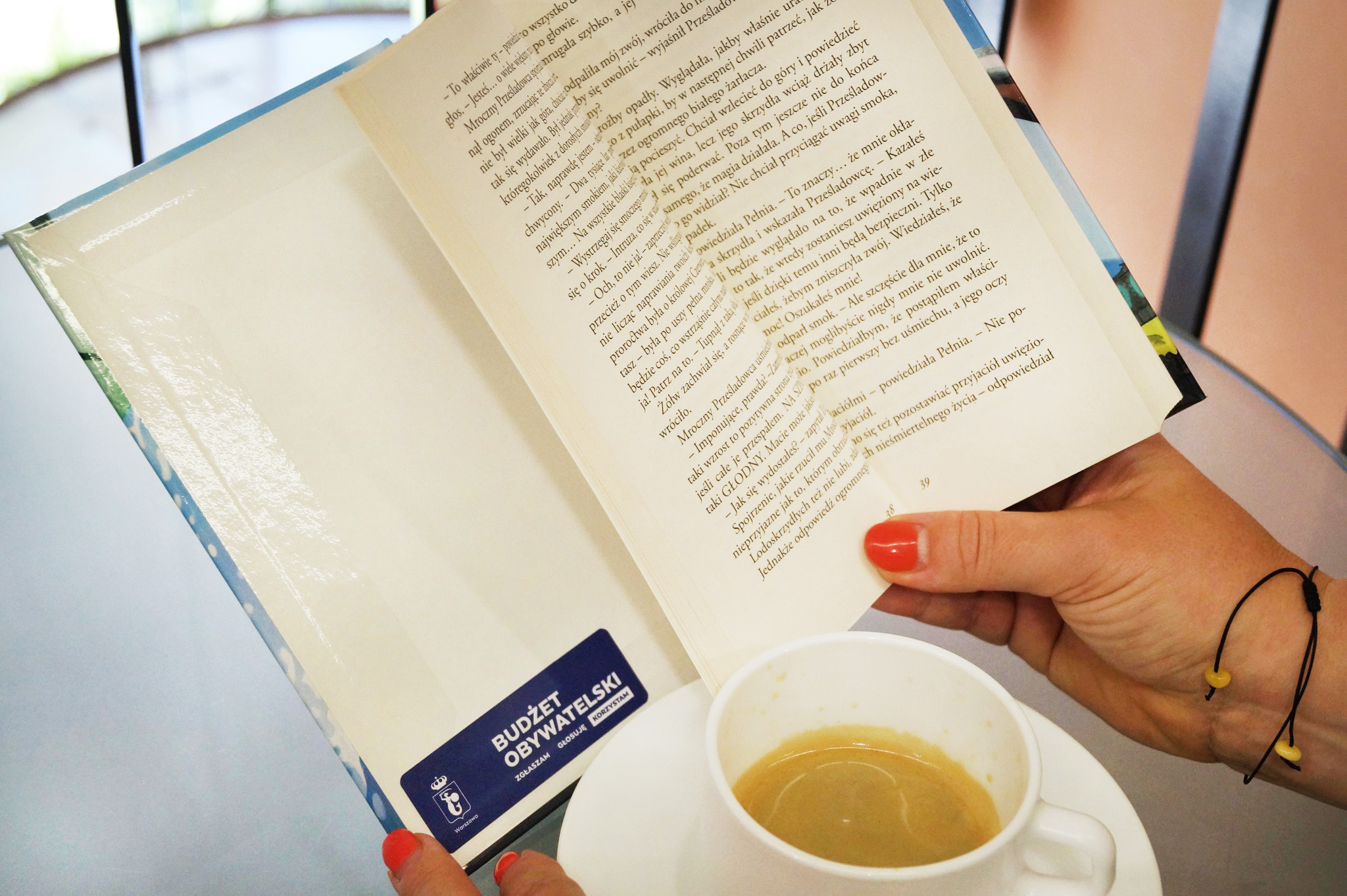Otwarta książka z naklejką Budżetu Obywatelskiego w kobiecej dłoni. Obok filiżanka kawy.