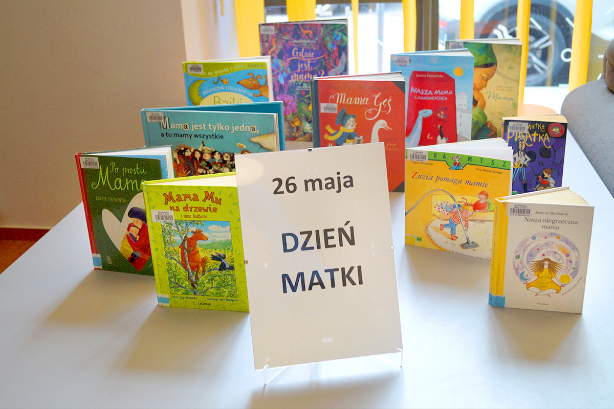 Wystawka kolorowych książek o mamach i dla mam ustawiona na stole niedaleko okna. Na pierwszym planie widać planszę z napisem: "26 maja. Dzień Matki". W tle wystawka różnych książek dla dzieci i mam.