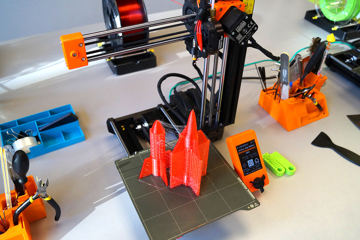 Drukarka 3D w trakcie pracy tworząca czerwony model rakiety. Widok z góry na stół warsztatowy