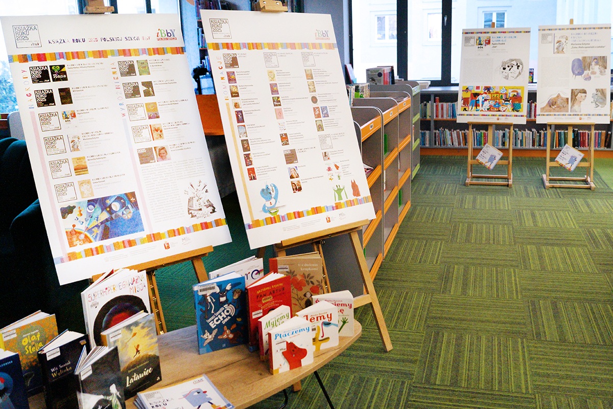 Po lewej dwie sztalugi z plakatami z wystawy, przed nimi stół z rozłożonymi nagrodzonymi książkami. W tle po prawej dwie sztalugi z plakatami z wystawy.
