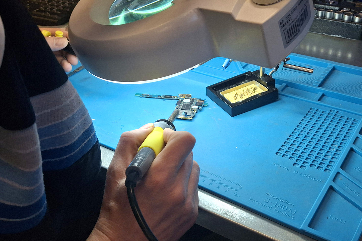 Widok zza pleców osoby lutującej mały element elektroniczny, leżący na błękitnej macie technicznej. Osoba korzysta z dużego szkła powiększającego. 