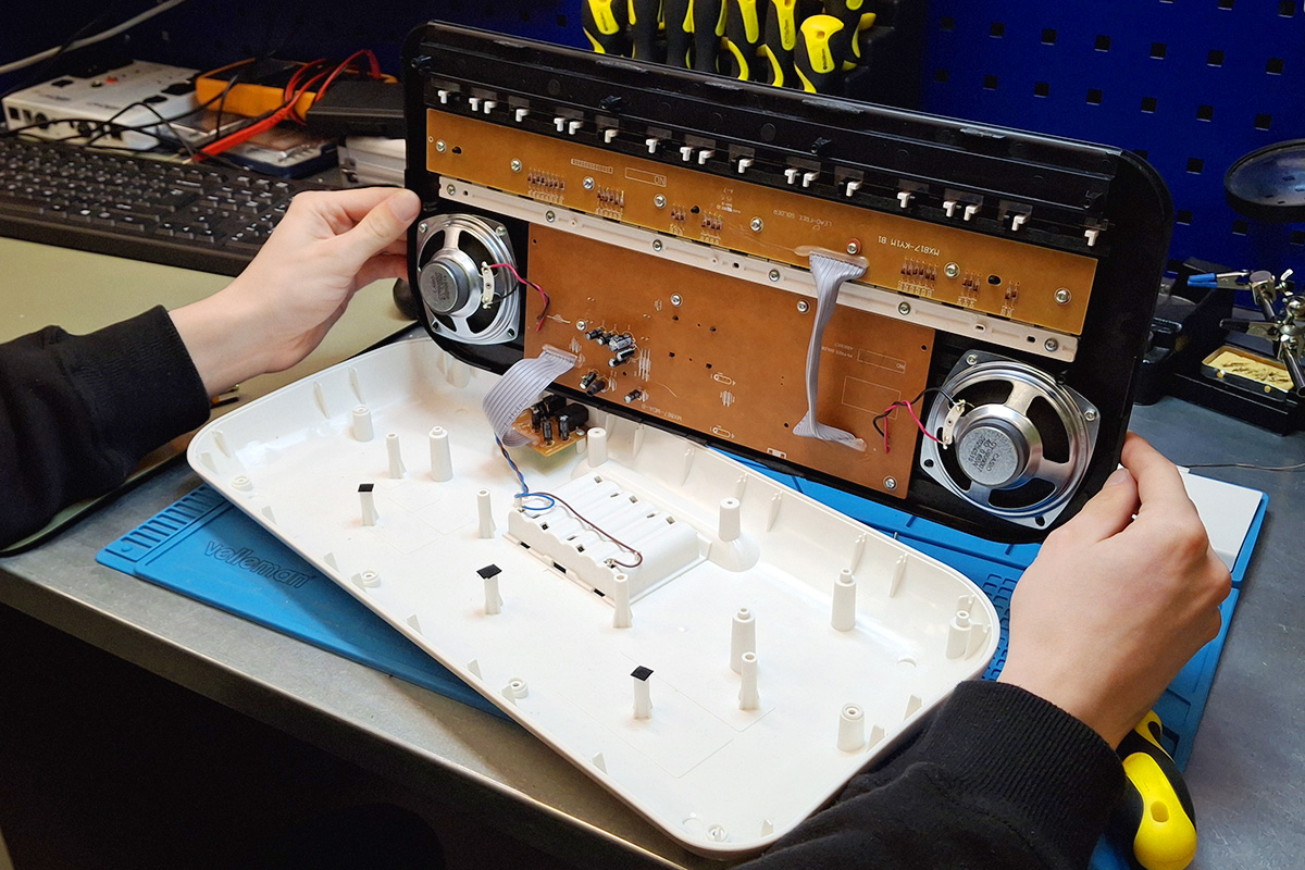 Ujęcie przedstawia wnętrze keyboarda ze zdjętą, białą obudową, trzymaną przez dwie ręce. W tle narzędzia i sprzęty na biurku warsztatowym - śrubokręty, klawiatura komputera, niebieska mata.