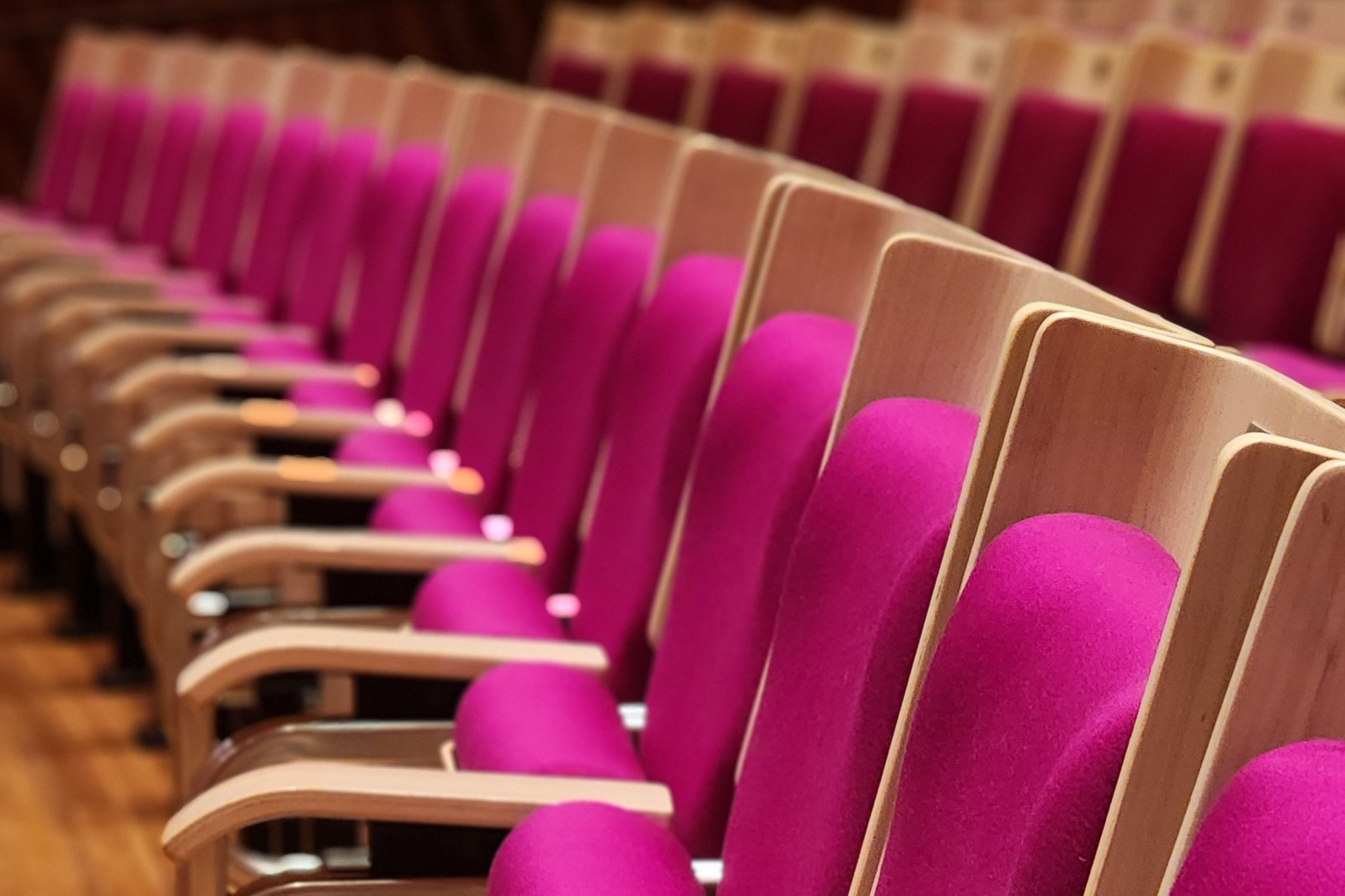 Zbliżenie na rzędy foteli sali kinowej w kolorze magenty z drewnianymi oparciami i podłokietnikami, ustawione na drewnianej podłodze.