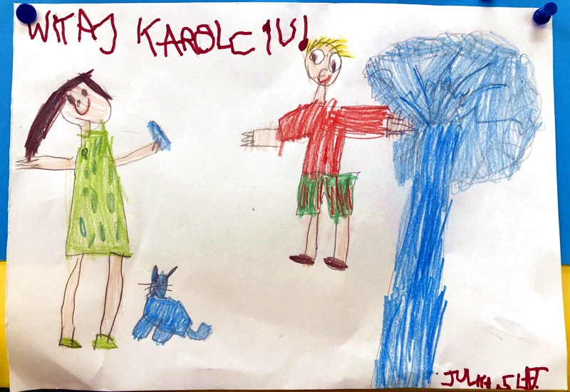 Dziecięcy rysunek. Na górze napis: Witaj Karolciu. Z prawej niebieskie drzewo. Od lewej uśmiechnięta kobieta w zielonej sukni rozpościera ręce.; niebieski kot oraz chłopiec w czerwonej bluzie i zielono-czerwonych szortach. Podpis: Julia, 5 lat.
