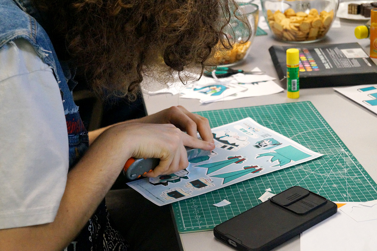 Widziana z profilu, młoda osoba z długimi, kręconymi, ciemnymi włosami wycina na macie technicznej papierowe elementy postaci Hatsune Miku. Dookoła i w tle na stole: kleje, kredki, przybory, ścinki papieru oraz miski z ciastkami i przekąskami.