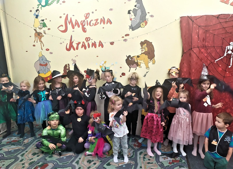 Gromada dzieci przebrana w stroje halloweenowe na tle żółtej ściany z obrazkami bajkowych postaci pozuje do zdjęcia.