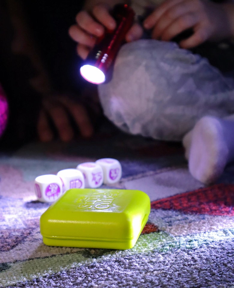 Na dywanie leży tajemnicze jasnozielone pudełko. Obok kostki z rysunkami od jakiejś gry. Dziecko oświetla obie rzeczy latarką.