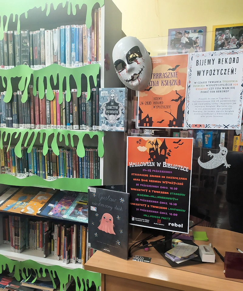 W prawej części stanowisko bibliotekarza z przyklejonymi różnymi halloweenowymi plakatami do pleksi. Z lewej regał z książkami i przyklejonymi zielonymi wycinankami jakby spływającej z półek mazi.