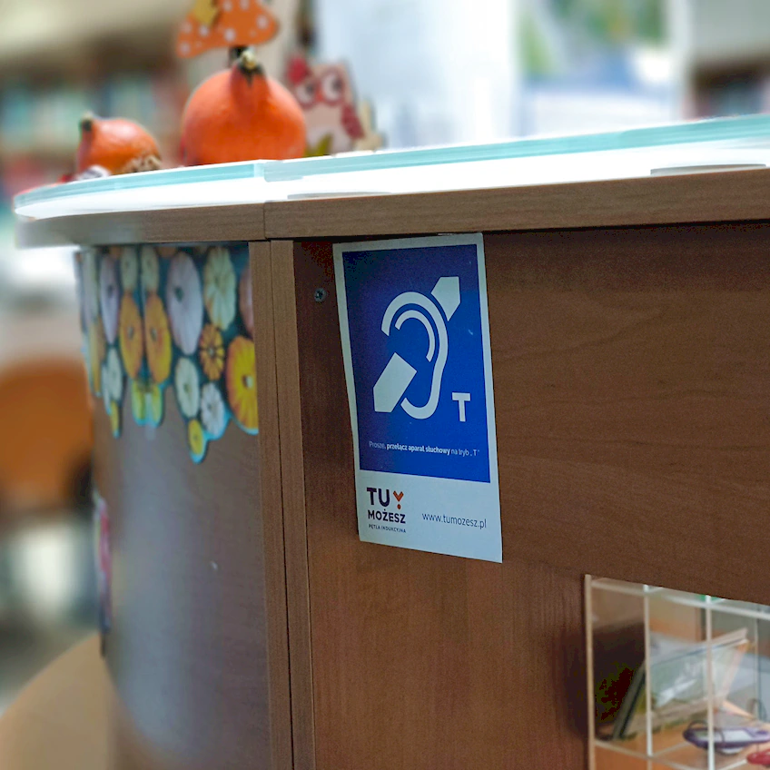 Zdjęcie fragmentu przedniej części lady bibliotecznej, na której naklejony jest znaczek informujący, że w tym miejscu jest zainstalowana pętla indukcyjna do aparatów słuchowych.