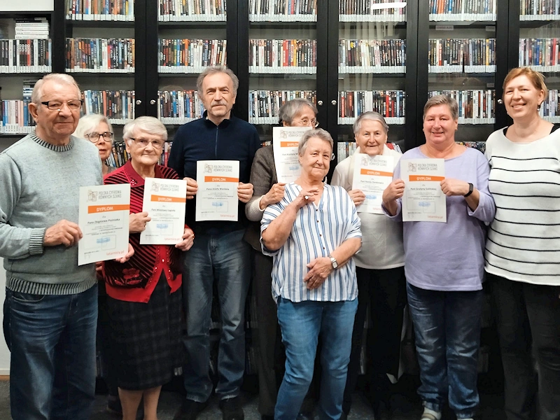 Uśmiechnięta grupa seniorów i seniorek trzyma w ręku dyplomy ukończenia kursu komputerowego. Na końcu z prawej uśmiecha się kierowniczka placówki. W tle pełne półki multimediów.