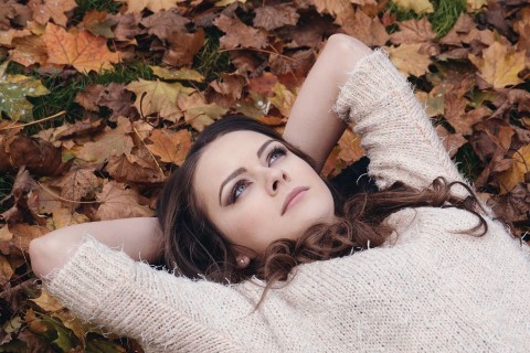 Młoda dziewczyna o ciemnych włosach leży na ziemi pośród liści. Ma na sobie biały sweter. Patrzy się przed siebie. Ma ręce splecione za głową.