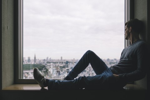 Młody mężczyzna siedzi w oknie, bokiem do obserwującego. Pada na niego cień. Patrzy się w dal, w kierunku niewyraźnej panoramy miasta.