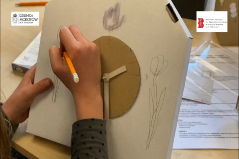 Ręka trzymająca ołówek rysuje tulipana na boku zegara zrobionego z kartonu.