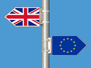 Na słupie dwa drogowskazy. Lewy prezentuje flagę Wielkiej Brytanii a prawy flagę Unii Europejskiej.