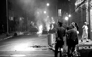 Zamieszanie na ulicy - policja, gapie, płonące śmietniki. Zdjęcie w tonacji czarno-białej.