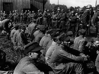 Żołnierze siedzący na ziemi, jeńcy z Westerplatte, pilnowani przez żołnierzy niemieckich z karabinami. 