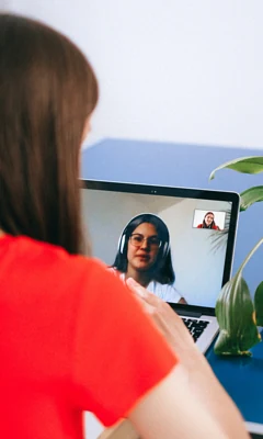 Odwrócona tyłem kobieta ubrana w czerwoną bluzkę rozmawia z inną kobietą widoczną na ekranie laptopa.