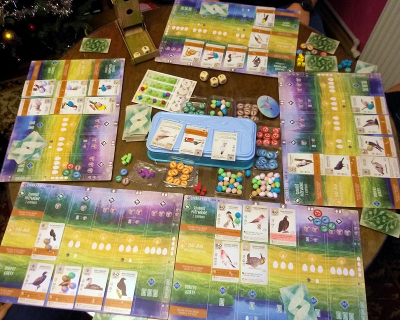 Na stole leży pięć kolorowych plansz graczy gry Na Skrzydłach. Na planszach leżą karty ptaków i kolorowe żetony. Na środku stołu, pomiędzy planszami leżą karty ptaków, żetony i jajka. Na górze po lewej stoi karmnik dla ptaków służący do rzucania kośćmi.