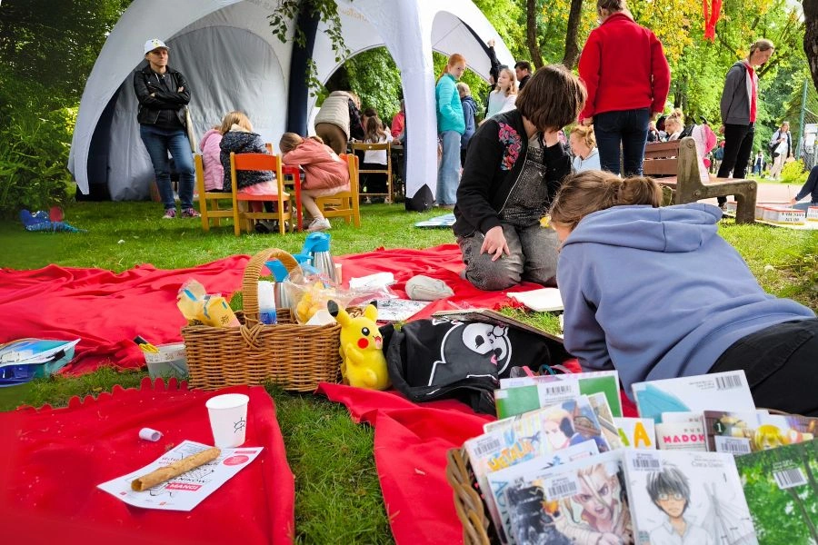 Piknik w parku, na kocach  osoby czytające książki, w tle namiot i uczestnicy imprezy