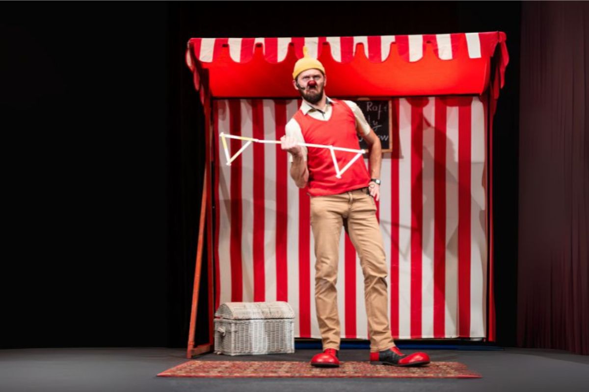 Klaun z czerwonym nosem trzyma w ręku rekwizyty teatralne